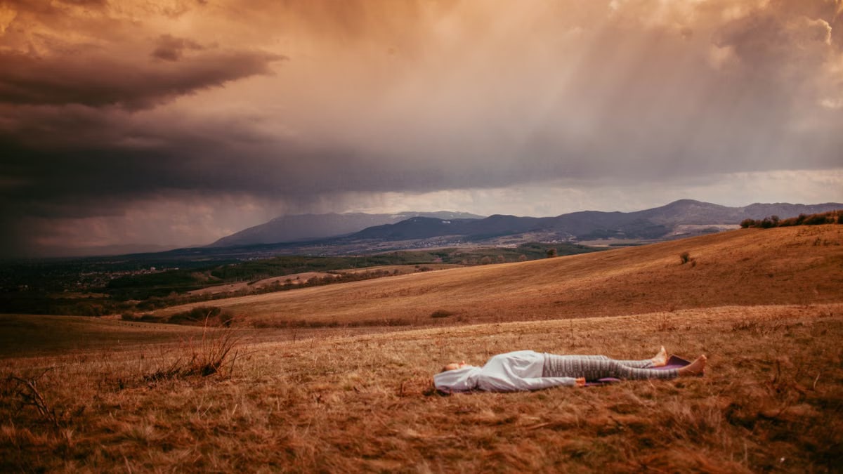 Individual lying on grassy field under a dramatic cloudy sky near mountains.